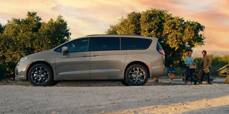 A family with a child is next to a gold minivan under a golden evening sky in an orchard.