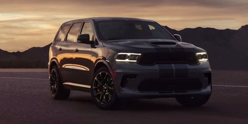 A dark gray Dodge Durango with racing stripes is parked on an empty road with mountains in the background at sunset.