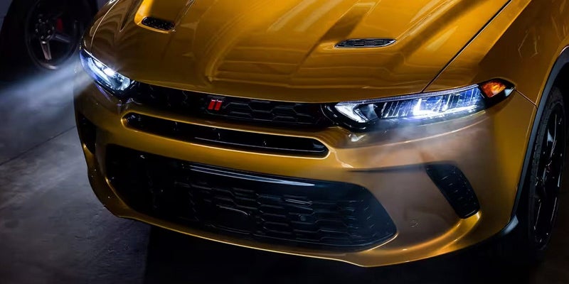 The front of a shiny gold Dodge vehicle with its headlights on, showcasing the grille and hood scoops.