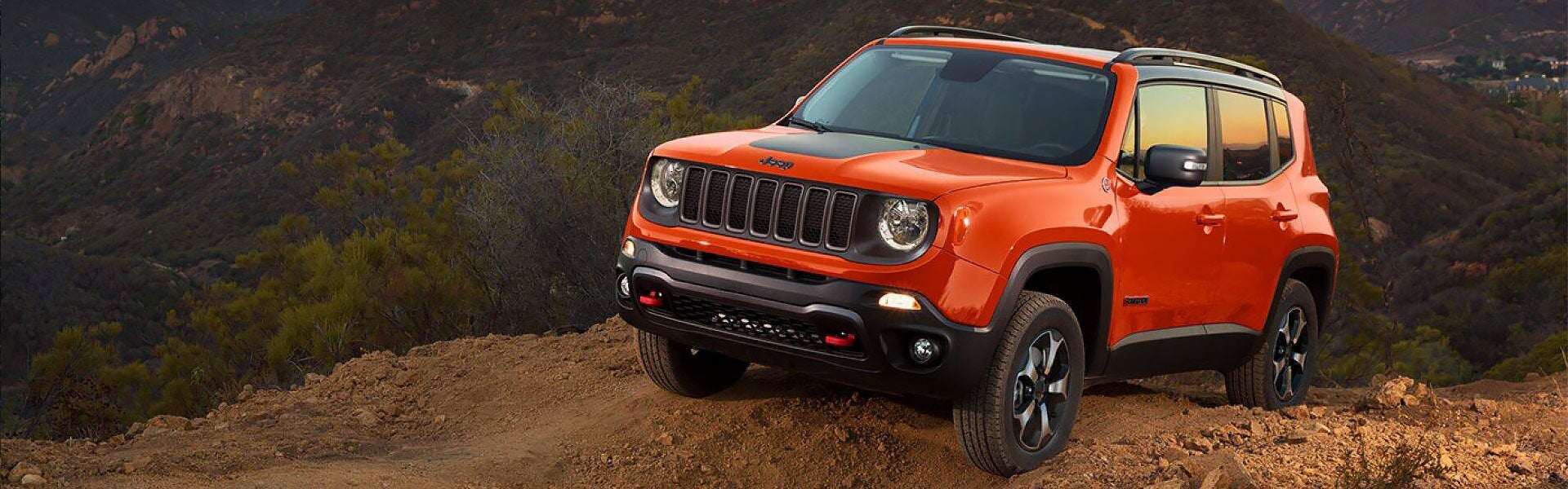 An orange Jeep Renegade with a black roof and hood stripe is parked on a dirt hill, facing slightly left.