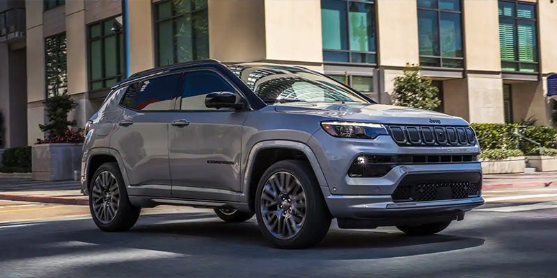 A silver Jeep Compass on a winding road with yellow wildflowers and rugged mountains under a blue sky.