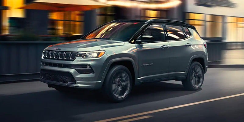 A grey Jeep Compass driving on a city street at dusk, with blurred background lights indicating motion.