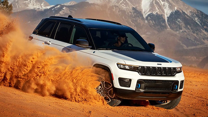 White Jeep Grand Cherokee 4xe kicking up sand on a desert slope, mountains in the background.