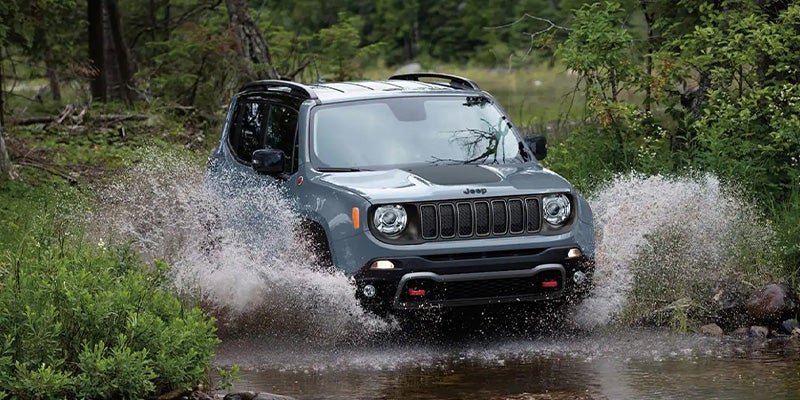 A grey Jeep drives through a shallow body of water, splashing water highly on each side, with a forest in the background.
