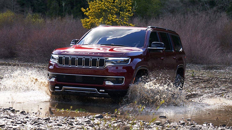 Red Jeep Wagoneer SUV drives through a shallow puddle on a dirt path, splashing water
