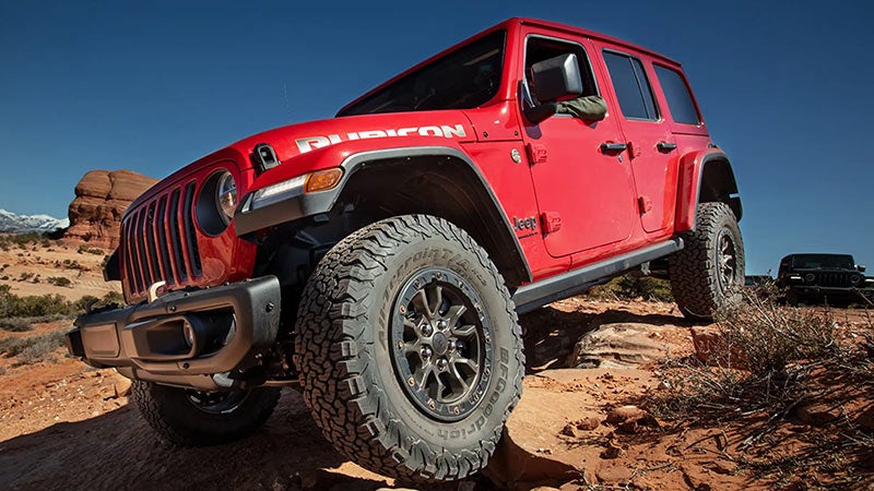 A red four-door Jeep Rubicon angles up a rocky dirt track in a desert landscape under a clear blue sky.