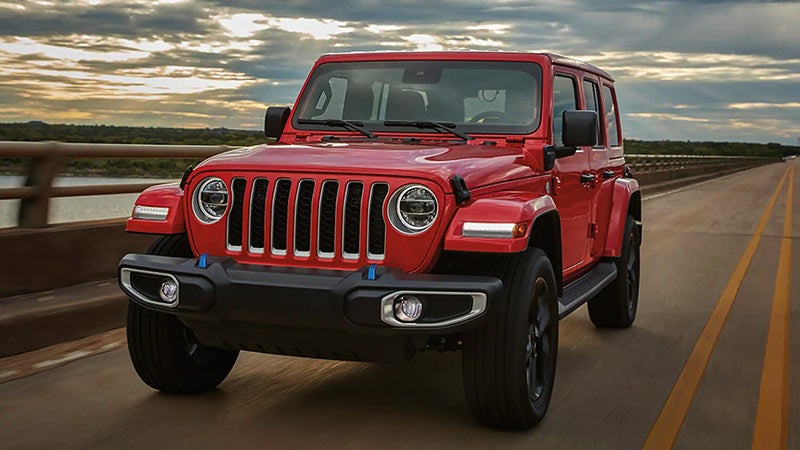 A red Jeep Wrangler drives on a bridge, facing slightly right. The sky is cloudy with a sunlit horizon.