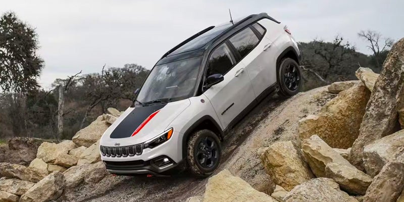 A white Jeep Compass SUV with a black hood stripe and red trim climbs a rocky incline.