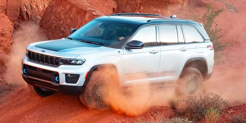 A white Jeep Grand Cherokee drives through reddish-brown dirt, kicking up rust-colored dust.