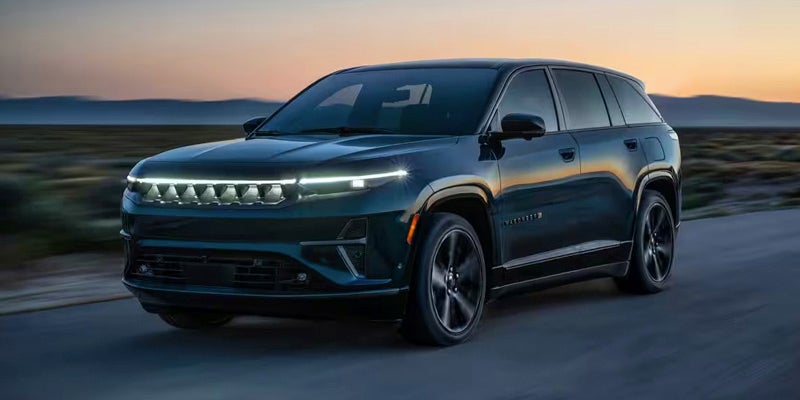 A sleek, dark green SUV with illuminated headlights drives on a road against a sunset sky.