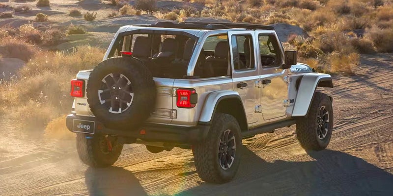 Rear view of a silver 4-door Jeep Wrangler with its roof removed, driving on a dirt road in a desert landscape.