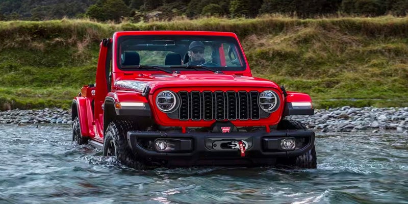 A red Jeep Wrangler drives through a shallow river with mountains and green land in the background.