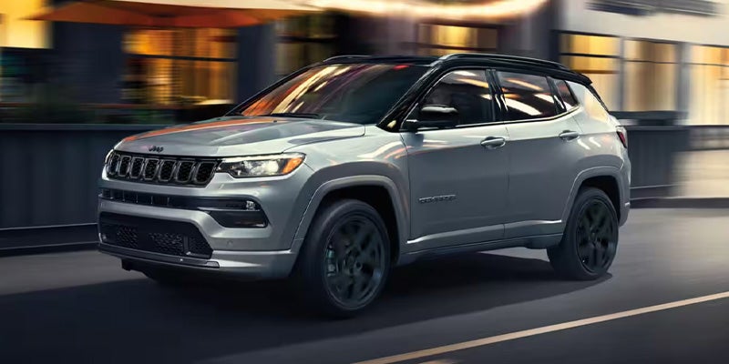 A light gray Jeep Compass Latitude with a black roof and black wheels drives along a dark street at dusk.