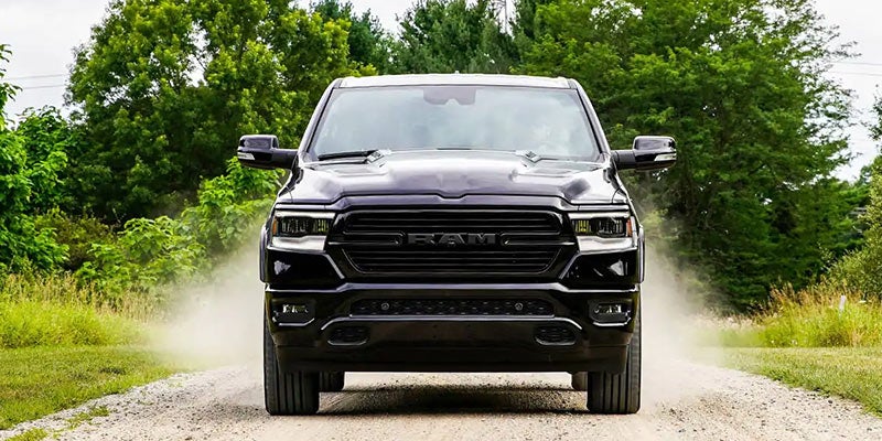Front view of a sleek black RAM truck driving on a dusty dirt road surrounded by lush green trees.