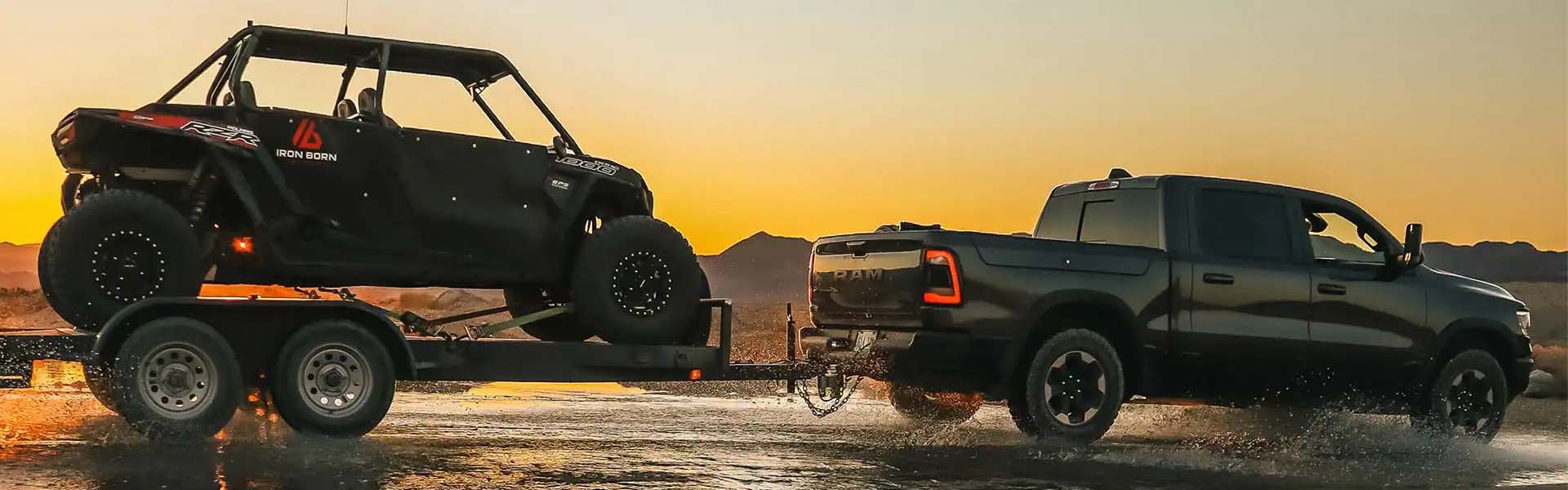 2022 Ram 1500 tows an RZR UTV on a trailer through shallow water at sunset, splashing water.