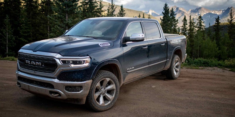 Dark blue Ram 1500 truck on a dirt road with mountains and pine trees in the background.