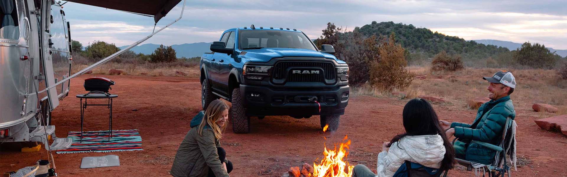 Ram 2500 and a camper trailer parked on reddish dirt with three people sitting around a campfire.