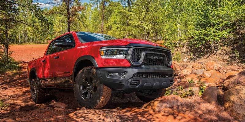 A red 2020 Ram Rebel truck drives over a rocky, dirt road in the woods.