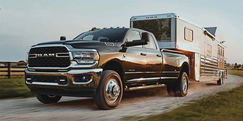 A black RAM dually pickup truck pulls a shiny silver horse trailer down a dirt road at sunset.