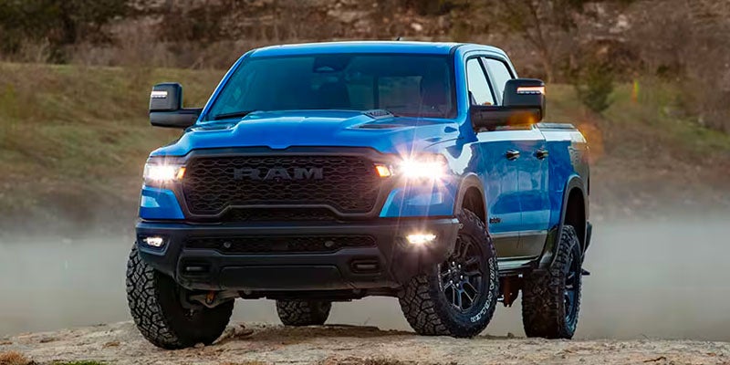 A bright blue RAM truck with its headlights on, driving on a dirt road.