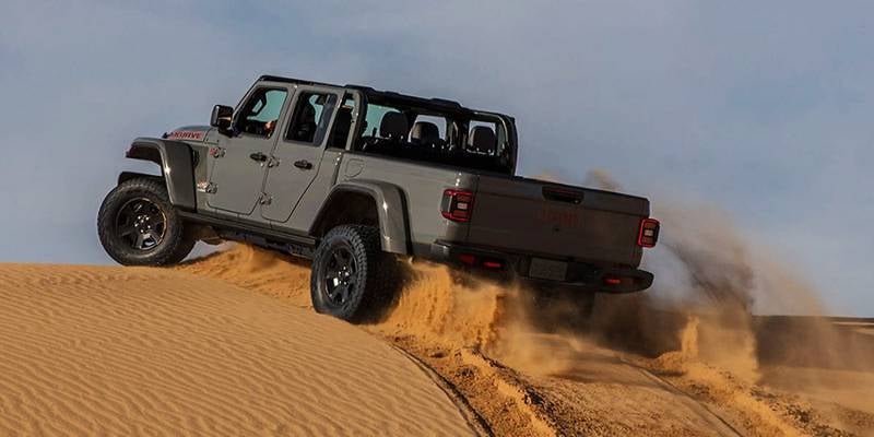 A gray pickup truck with an open top drives up a sandy dune, kicking up sand behind it.